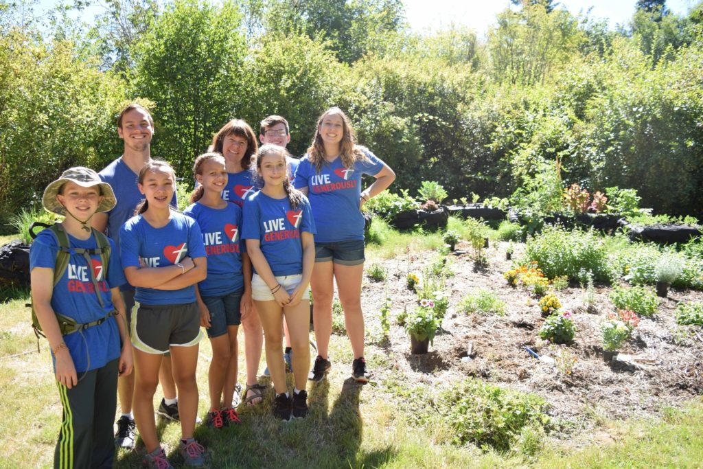 A group of adults and childen wearing shirts that say "Live Generously" standing next to the garden they volunteered to clean up at Camp Lutherwood Oregon